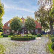 Blick auf ein altes Gutshaus mit rotem Dach und roten Backsteinen im Herbst. Davor sind zwei Bäumen angeordnet, die Sonne scheint von rechts oben durch eine alte Baumkrone und ein runder, bepflanzter Platz ist im Vordergrund zu sehen.