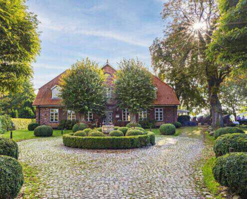 Blick auf ein altes Gutshaus mit rotem Dach und roten Backsteinen im Herbst. Davor sind zwei Bäumen angeordnet, die Sonne scheint von rechts oben durch eine alte Baumkrone und ein runder, bepflanzter Platz ist im Vordergrund zu sehen.