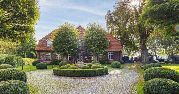 Blick auf ein altes Gutshaus mit rotem Dach und roten Backsteinen im Herbst. Davor sind zwei Bäumen angeordnet, die Sonne scheint von rechts oben durch eine alte Baumkrone und ein runder, bepflanzter Platz ist im Vordergrund zu sehen.