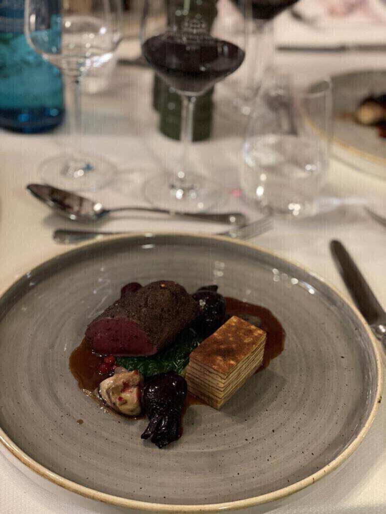 Auf einem grauen Teller liegt ein Hauptgericht aus Fleisch, Kartoffeln und Rüben. Im Hintergrund sind Wein- und Wassergläser sowie Besteck zu sehen.