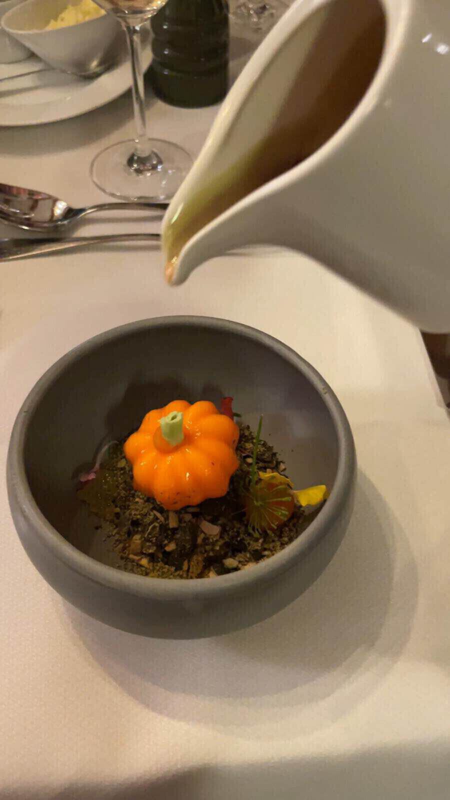 Ein kleiner orangefarbener Kürbis ist in der Mitte einer kleinen tiefen Schale auf Gemüse gelegt worden und wird nun mit einer Gemüsesauce übergossen.