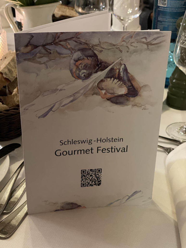 Eine Speisekarte mit einem gemahlenen Muschelbild, einem QR-Code und dem Namen "Schleswig-Holstein Gourmet Festival" steht auf einem gedeckten Tisch.