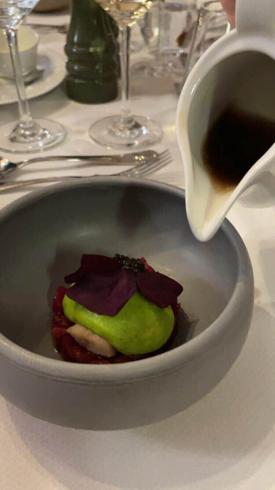 Ein Gericht mit Fleisch und Fisch wird in einer kleinen tiefen Schale mit einer Fleischbrühe am Tisch übergossen.