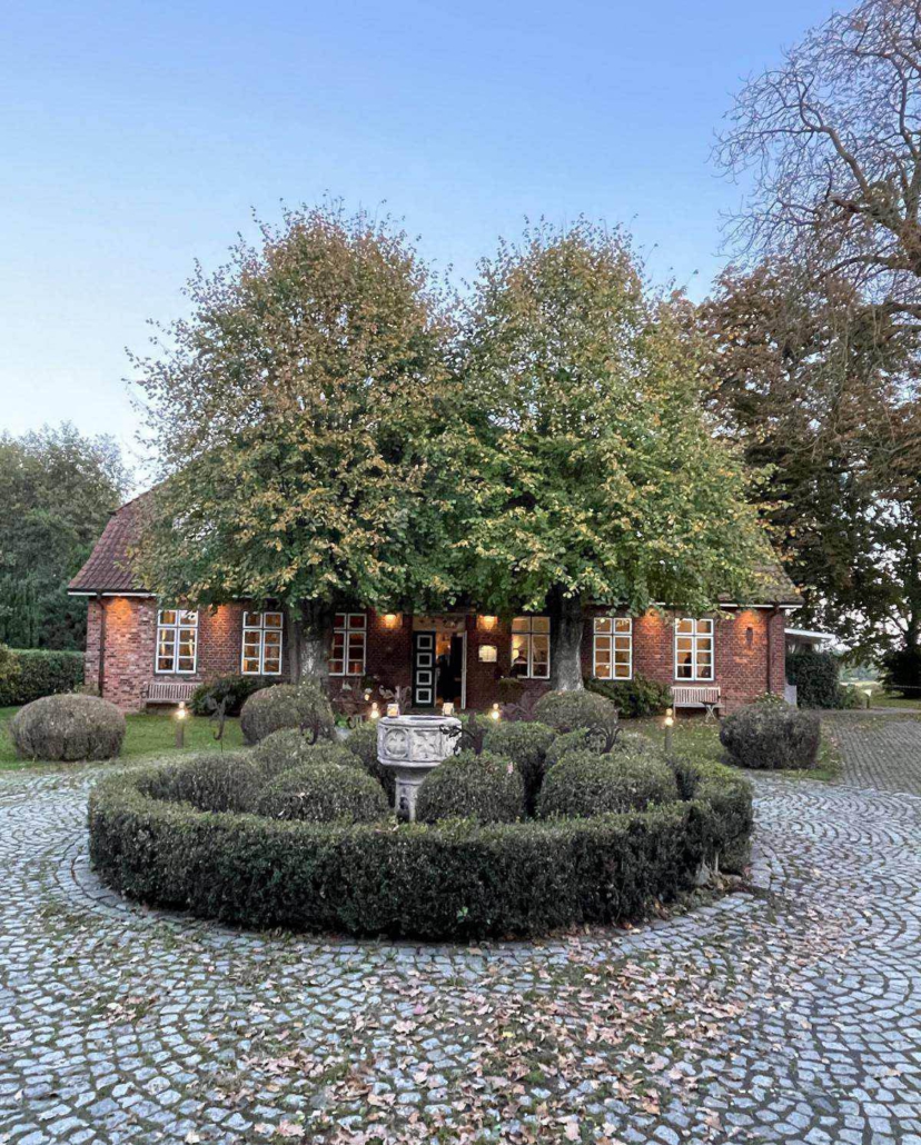 Blick auf ein altes Gutshaus aus rotem Backstein, mit zwei Bäumen und einem kreisförmigen Platz davor.