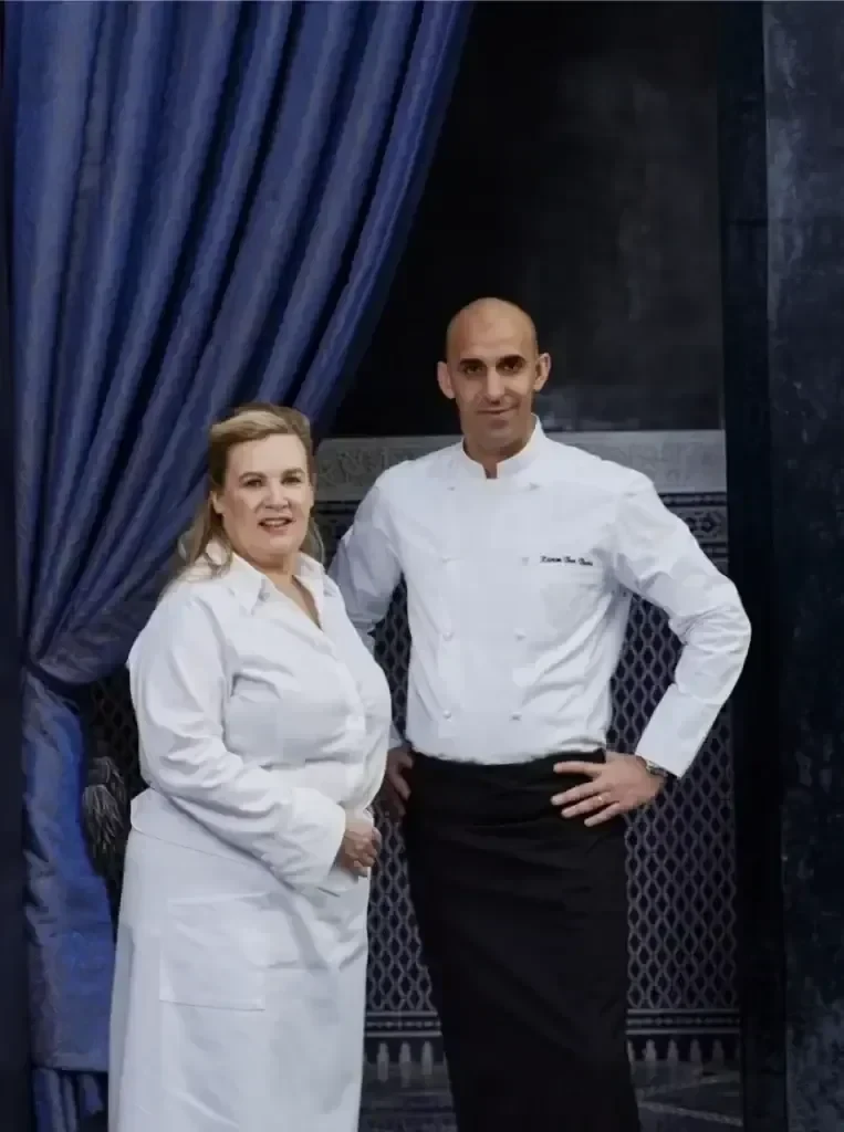 Zwei Köche in weißen Jacken stehen nebeneinander vor einem blauen Vorhang und einer gemusterten Wand und schauen in die Kamera.