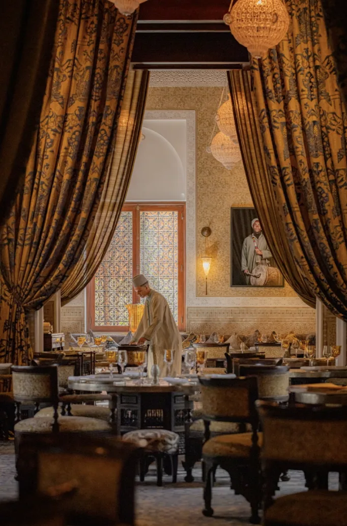 Eine Person in traditioneller Kleidung deckt den Tisch in einem verzierten, warm beleuchteten Esszimmer mit gemusterten Vorhängen, Plüschstühlen und einem Porträt an der Wand.