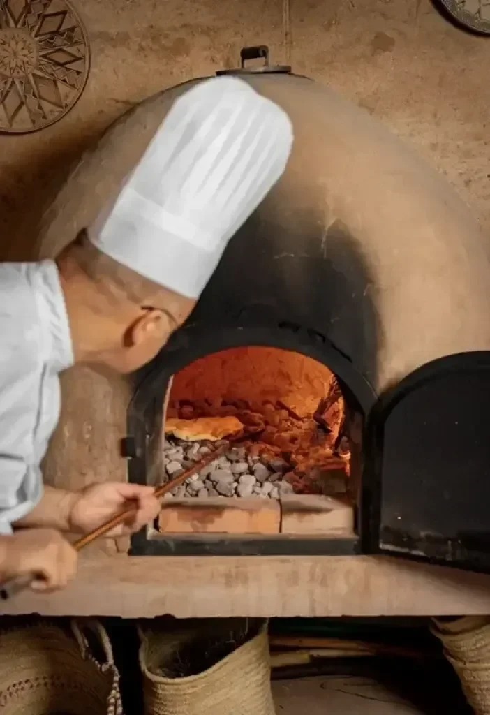 Ein Koch in weißer Uniform und Mütze legt die Speisen in einen traditionellen, mit heißen Kohlen gefüllten Lehmofen.