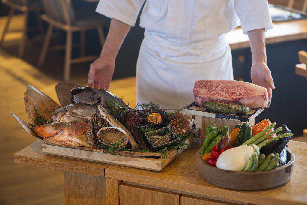 Ein Koch steht hinter Tabletts mit frischem Fisch, Meeresfrüchten, Gemüse und einem großen Stück marmorierten Rindfleisch auf einem Tresen in einem Restaurant.