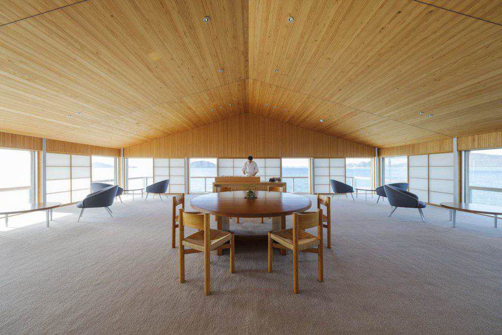 Geräumiges Zimmer mit Holzdecke, rundem Esstisch und Stühlen in der Mitte, Sesseln an großen Fenstern mit Meerblick, Person am Schreibtisch.