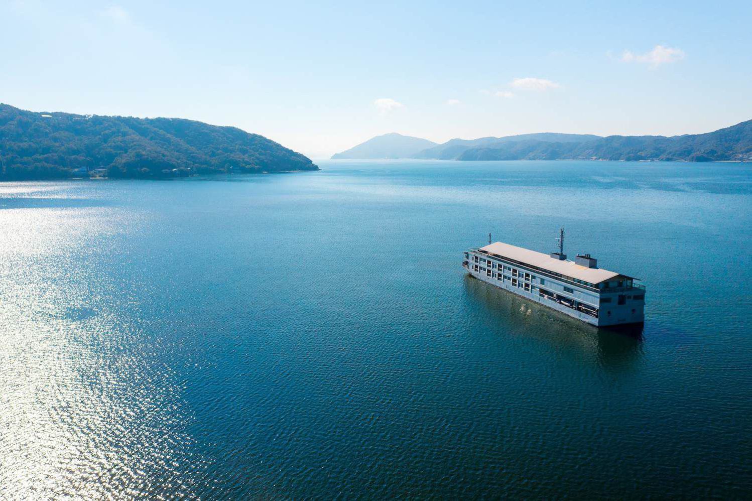 Ein großes Hausboot schwimmt auf ruhigem, blauem Wasser, umgeben von weit entfernten bewaldeten Hügeln unter einem klaren Himmel.