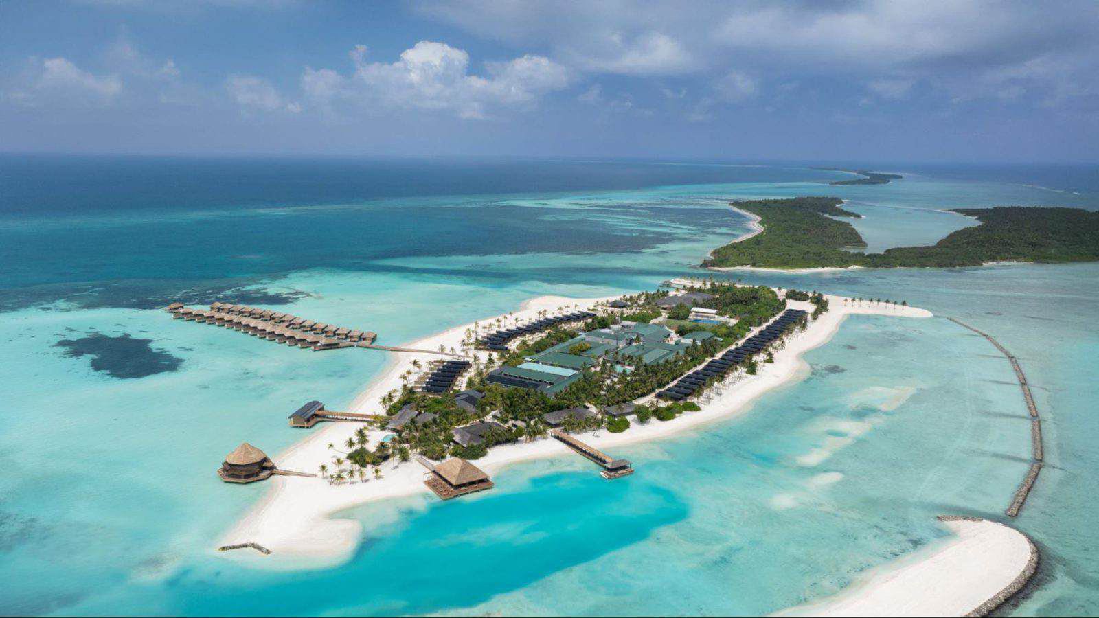 Luftaufnahme eines tropischen Inselresorts, umgeben von türkisfarbenem Meer, weißen Sandstränden, Überwasserbungalows und üppigem Grün mit angrenzenden kleinen Inseln.