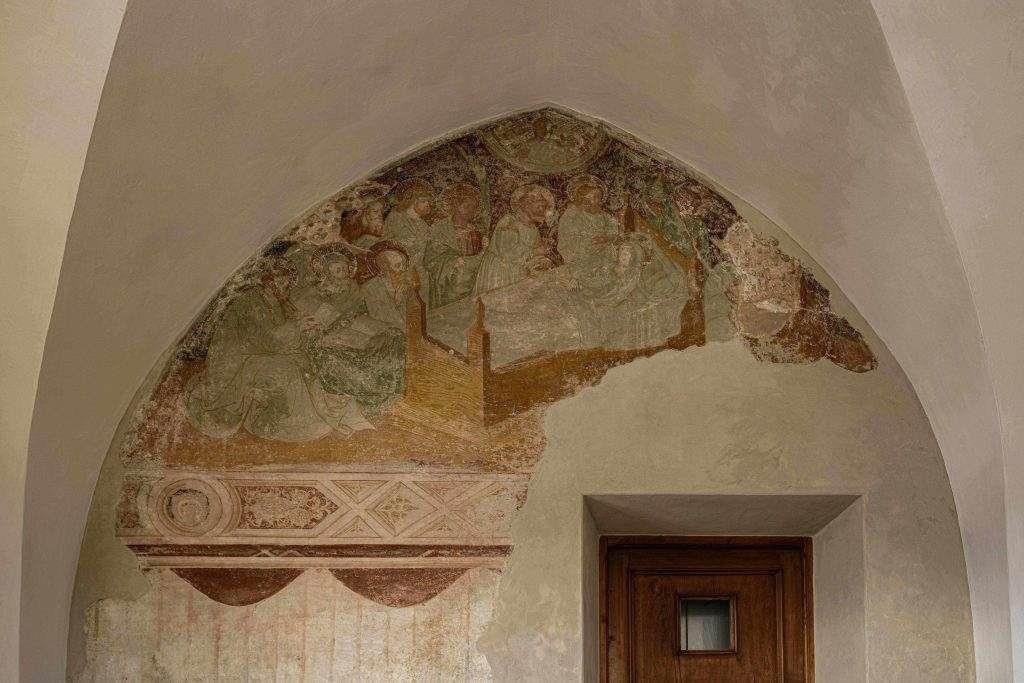 Detail eines gothischen Freskos an einer gewölbten Wand über einer Tür, das religiöse Figuren in einem historischen Kontext darstellt.