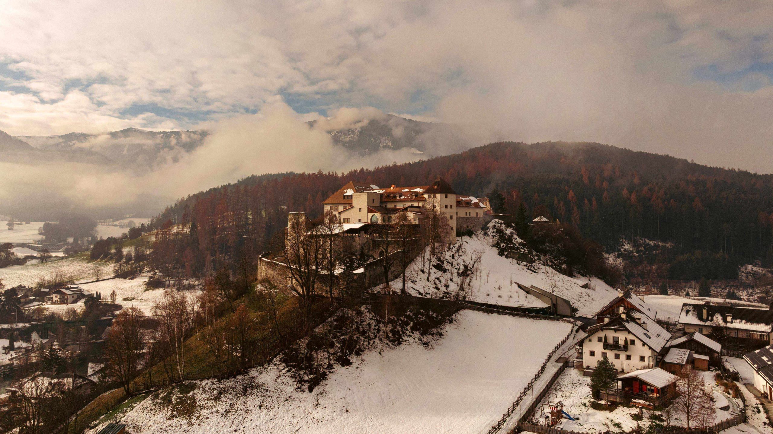Eine Luftaufnahme eines großen, befestigten Schlosses auf einem Hügel, das von tiefem, schneebedecktem Wald und einem kleinen Dorf am Fuße des Hügels umgeben ist, unter einem teilweise bewölkten Himmel.