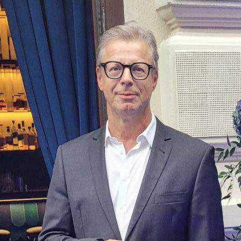 Ein Mann mittleren Alters, der eine Brille und einen grauen Anzug trägt, steht drinnen in der Nähe eines blauen Vorhangs und eines Barbereichs.