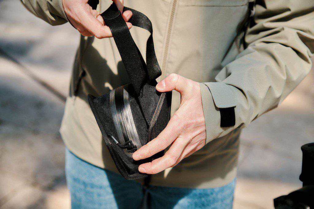 Eine Person in heller Jacke und Jeans öffnet eine kleine schwarze Tasche mit Reißverschluss und einem Griff, der das FIDLOCK-System im Freien zeigt.