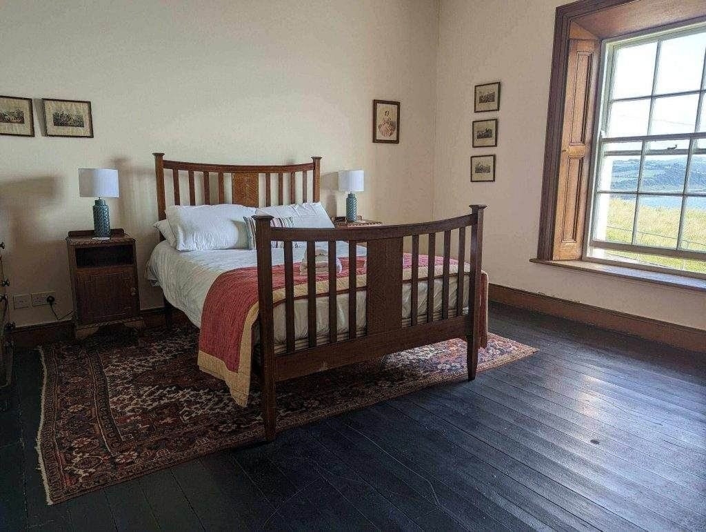 Ein Holzbett mit weißer Bettwäsche und einer roten Decke steht auf einem Orientteppich in einem Raum mit dunklem Holzboden, einem Fenster und gerahmten Bildern an cremefarbenen Wänden.