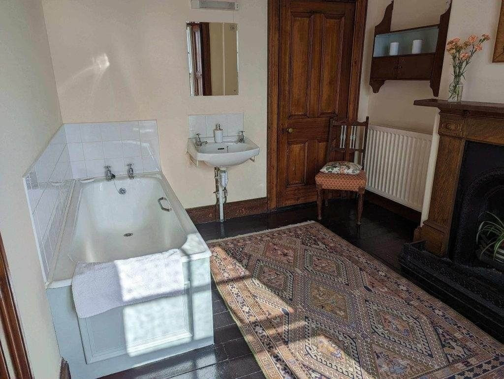 Ein Badezimmer mit einer weißen Badewanne, einem an der Wand befestigten Waschbecken, einem Holzstuhl mit Kissen, einem gemusterten Teppich, einem Kamin und einem Spiegel über dem Waschbecken.