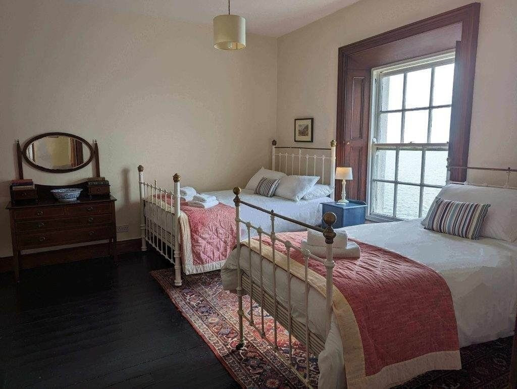 Zwei Einzelbetten mit weißen Metallrahmen und rosa Bezügen stehen in einem Schlafzimmer mit einem großen Fenster, einer Holzkommode, einem Spiegel und einem Teppich auf einem dunklen Holzboden.