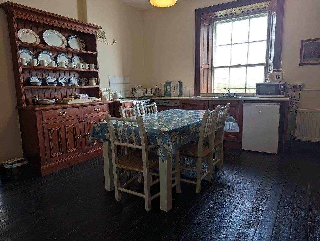 Eine Küche mit einem hölzernen Esstisch, der mit einem blauen Tischtuch bedeckt ist, weißen Stühlen, einem hölzernen Schrank mit Geschirr, einem Fenster, einem Kühlschrank, einer Mikrowelle und einem Wasserkocher auf dunklem Holzboden.