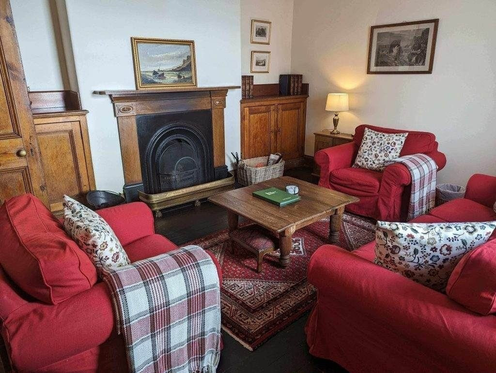 Ein gemütliches Wohnzimmer mit roten Sesseln und Sofa, Couchtisch aus Holz, Kamin, gemustertem Teppich und karierten Decken, dekoriert mit gerahmten Bildern und einer Tischlampe.