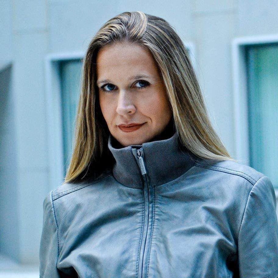 Eine Frau mit langen, glatten blonden Haaren trägt eine graue Jacke mit Reißverschluss und steht vor einem Gebäude mit hellblauen Fenstern.