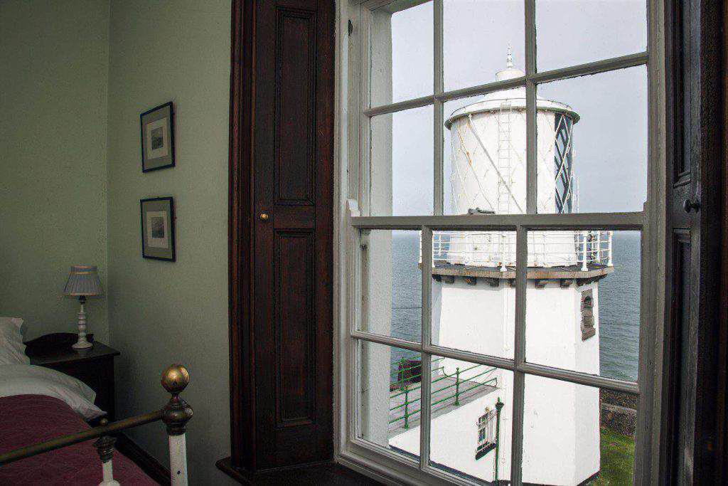 Ein Leuchtturm mit reicher maritimer Geschichte ist durch ein großes Fenster von einem Schlafzimmer in Irland aus zu sehen, wobei gerahmte Bilder, eine Lampe und ein Bett teilweise sichtbar sind - perfekt für eine unvergessliche Übernachtung im Leuchtturm.