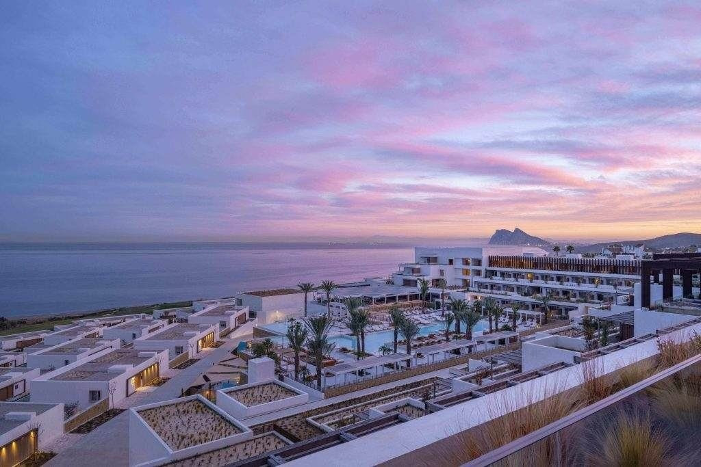 Modernes Resort am Meer mit weißen Gebäuden, Palmen und einem zentralen Pool, mit Blick auf das Meer bei Sonnenuntergang und einen felsigen Berg in der Ferne.