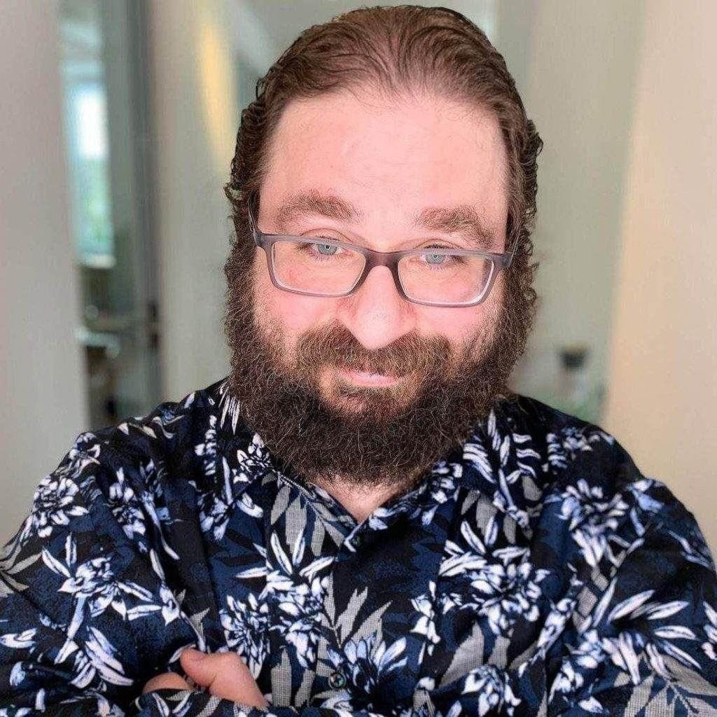 Ein Mann mit Brille, Bart und langen Haaren steht in einem dunklen geblümten Hemd im Haus und schaut mit verschränkten Armen in die Kamera.