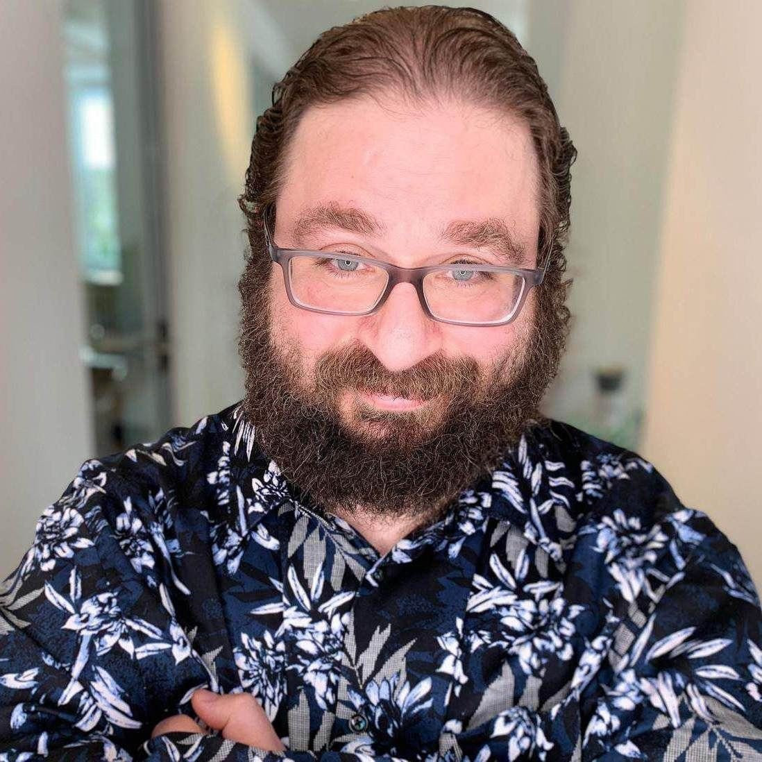 Ein Mann mit Brille, Bart und langen Haaren steht in einem dunklen geblümten Hemd im Haus und schaut mit verschränkten Armen in die Kamera.