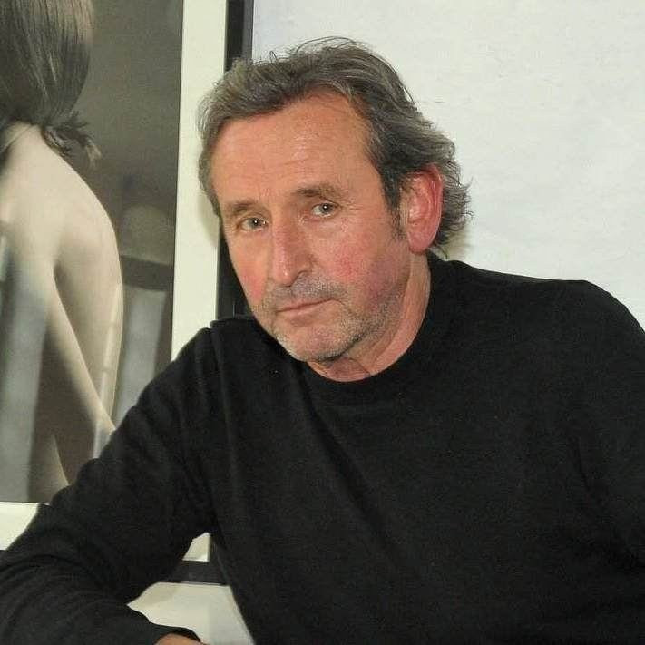 Ein Mann mittleren Alters mit grauem Haar und schwarzem Pullover sitzt in einem Haus vor einem gerahmten Schwarz-Weiß-Foto.