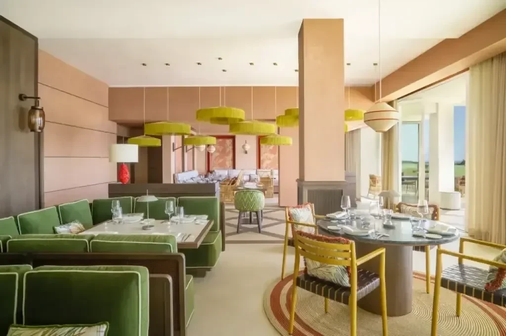 Modernes Restaurantinterieur im La Hacienda Links Golf Resort mit grünen Samtbestuhlungen, runden Tischen zum Essen, gelb-grünen Hängeleuchten und großen Fenstern, die natürliches Licht in dieses Top 100 Golfresort lassen.
