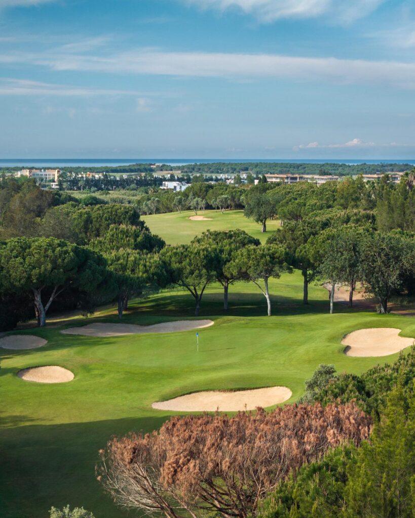 Ein weitläufiger, sonnenbeschienener Golfplatz mit Blick auf das Meer. Im Vordergrund liegt ein gepflegtes grünes Putting-Green mit einer blauen Fahne, umgeben von mehreren hellen Sandbunkern. Dahinter erstreckt sich ein dichter Pinienhain. In der Ferne sind weiße Küstengebäude und der blaue Horizont des Ozeans unter einem leicht bewölkten Himmel zu sehen.