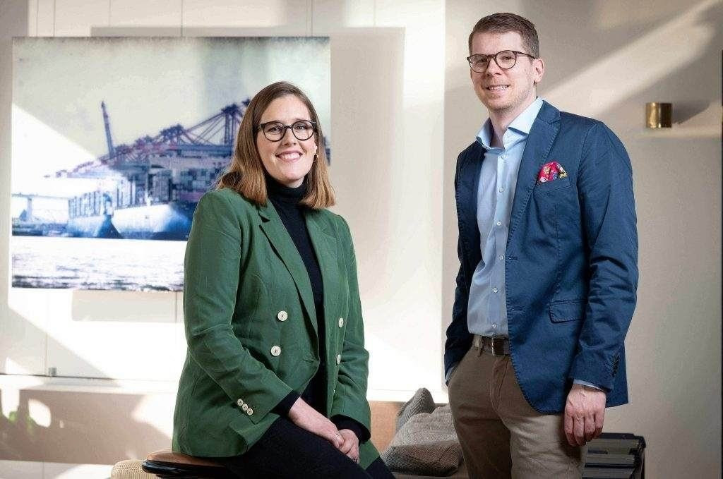 Annie Dörfelt und Henrik Maaß, die Gründer von Liquid Grape, lächeln in die Kamera. Annie sitzt links in einem grünen Blazer, Henrik steht rechts daneben in einem blauen Sakko mit buntem Einstecktuch. Im Hintergrund hängt ein großformatiges Bild eines Containerschiffs.