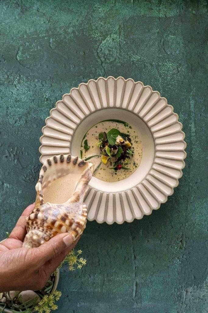 Eine Hand gießt Soße aus einer Muschelschale in eine dekorative Schale, die ein Gourmetgericht mit Kräutern und Gemüse auf einer grünen Fläche enthält.