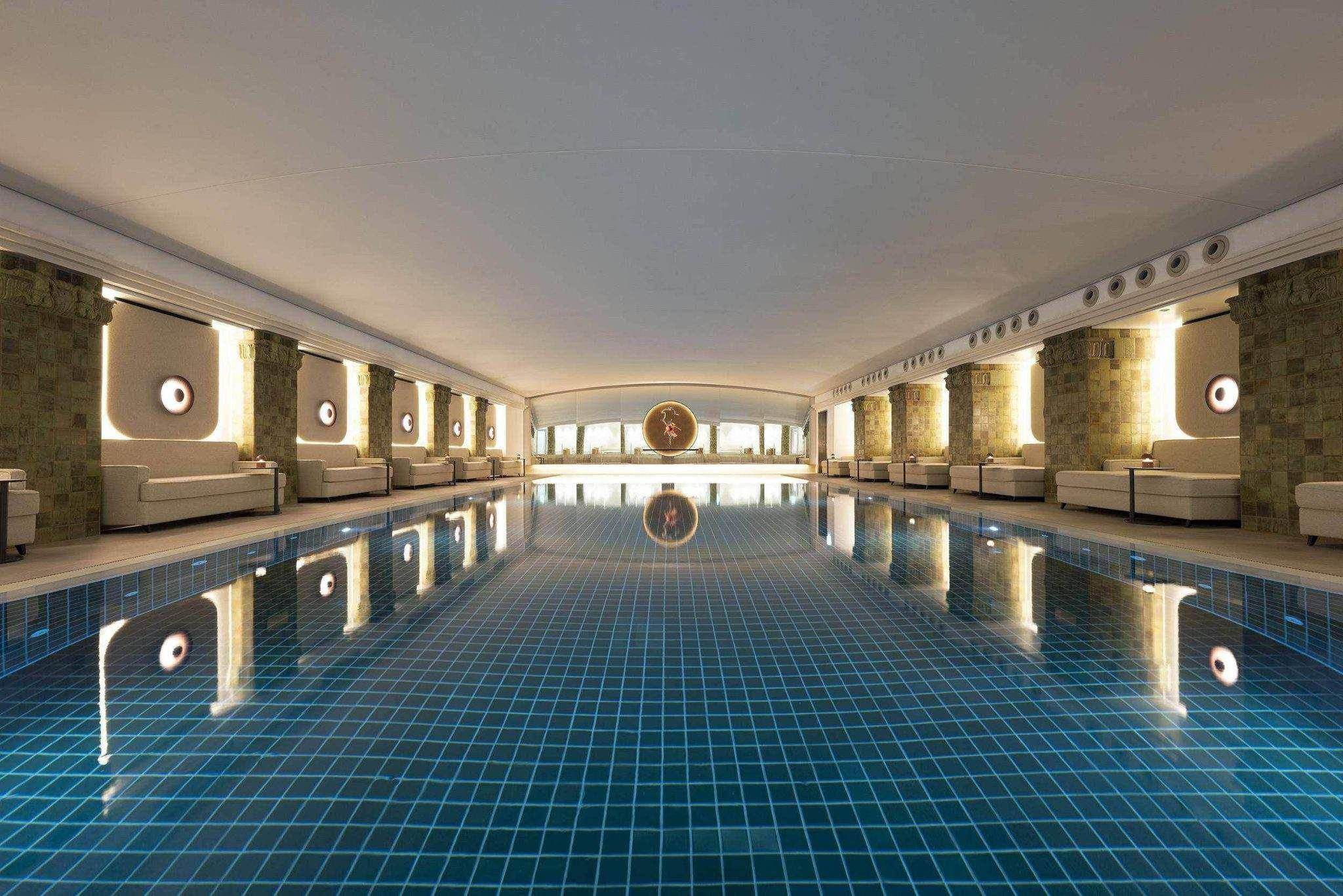 Großzügiger, beleuchteter Innenpool mit blauen Fliesen und symmetrischen Lounge-Sitzbereichen im Levante Spa des Conrad Hamburg.