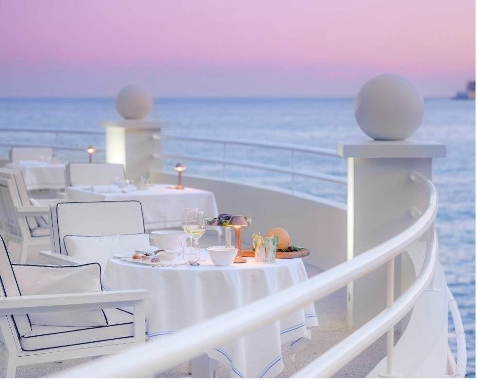 Elegante Außenterrasse eines Gourmet-Restaurants in Monaco bei Dämmerung mit gedeckten Tischen, weißen Stühlen mit dunkelblauen Paspeln und Blick auf das Mittelmeer unter einem rosa-blauen Himmel.
