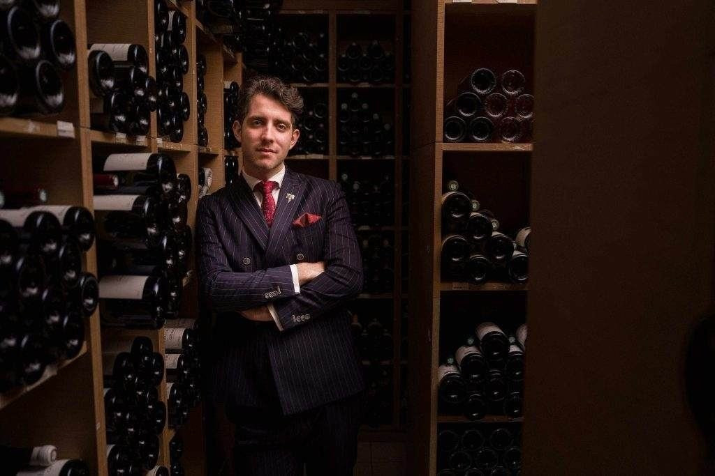 Ein Porträt von Francesco Cosci in einem dunklen Nadelstreifenanzug mit roter Krawatte. Er steht mit verschränkten Armen in einem stimmungsvoll beleuchteten Weinkeller, umgeben von Regalen voller Weinflaschen.