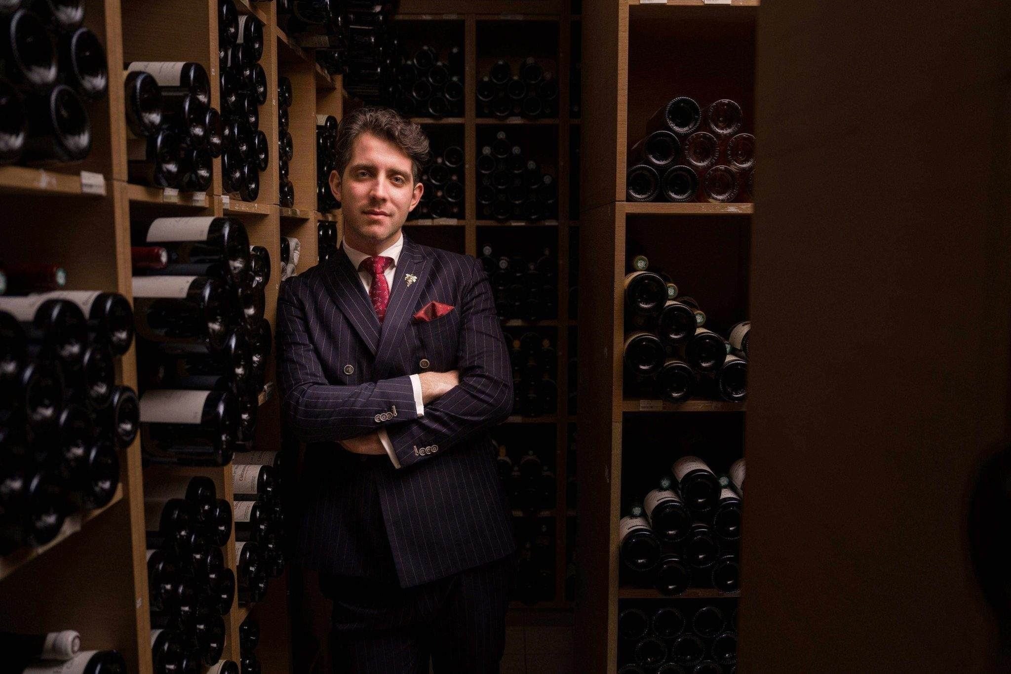 Ein Porträt von Francesco Cosci in einem dunklen Nadelstreifenanzug mit roter Krawatte. Er steht mit verschränkten Armen in einem stimmungsvoll beleuchteten Weinkeller, umgeben von Regalen voller Weinflaschen.