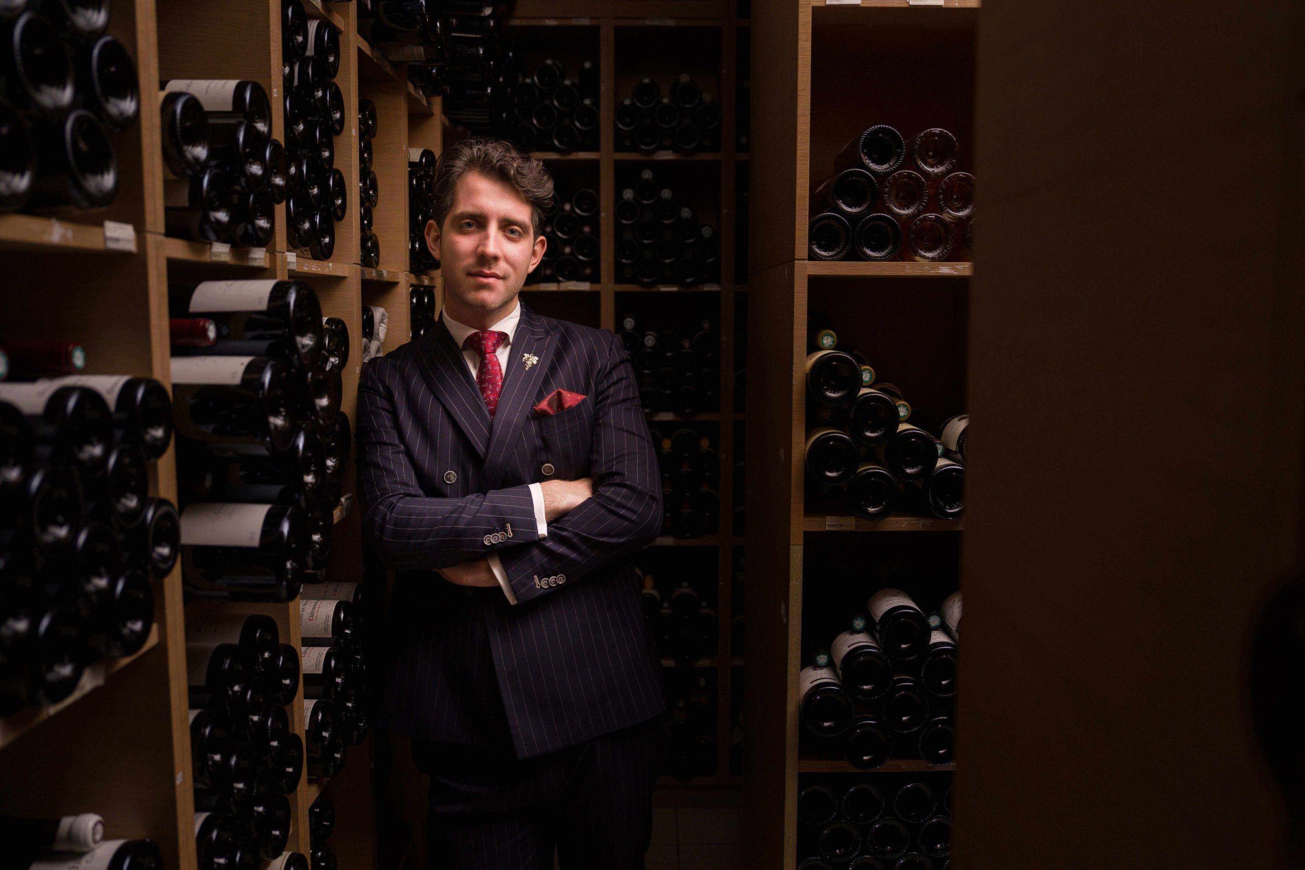 Ein Porträt von Francesco Cosci in einem dunklen Nadelstreifenanzug mit roter Krawatte. Er steht mit verschränkten Armen in einem stimmungsvoll beleuchteten Weinkeller, umgeben von Regalen voller Weinflaschen.