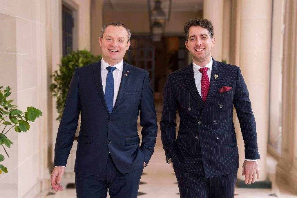 Zwei lächelnde Männer in eleganten Business-Anzügen, Frédéric Kaiser und Francesco Cosci, gehen nebeneinander durch einen hellen, luxuriösen Hotelflur mit hohen Decken und Säulen.