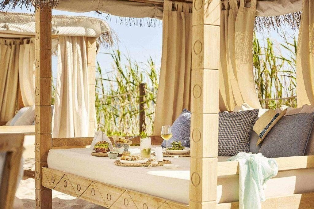 Ein gedeckter Tisch mit Sushi, einem Burger, Pommes und Drinks auf einem gemütlichen Cabana-Bett aus Holz mit hellen Vorhängen und Blick ins Grüne.