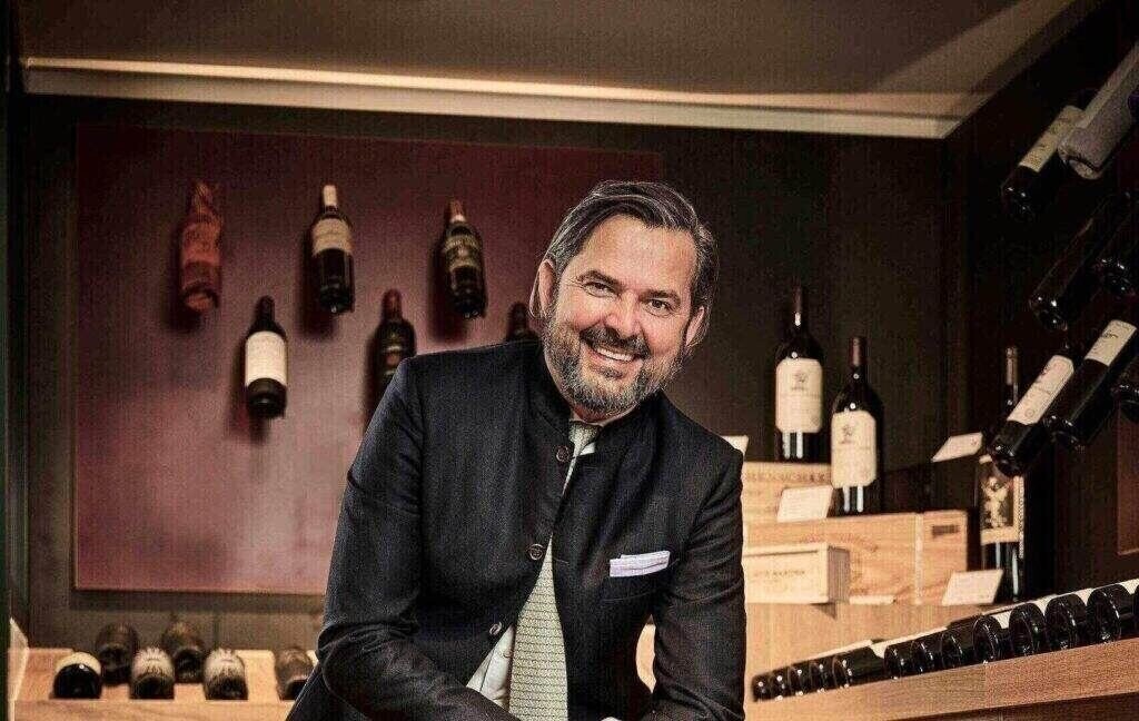 Ein Mann in dunkler Jacke und Krawatte lächelt in einem Weinkeller, umgeben von Mövenpick-Weinflaschen in Regalen und Holzkisten.