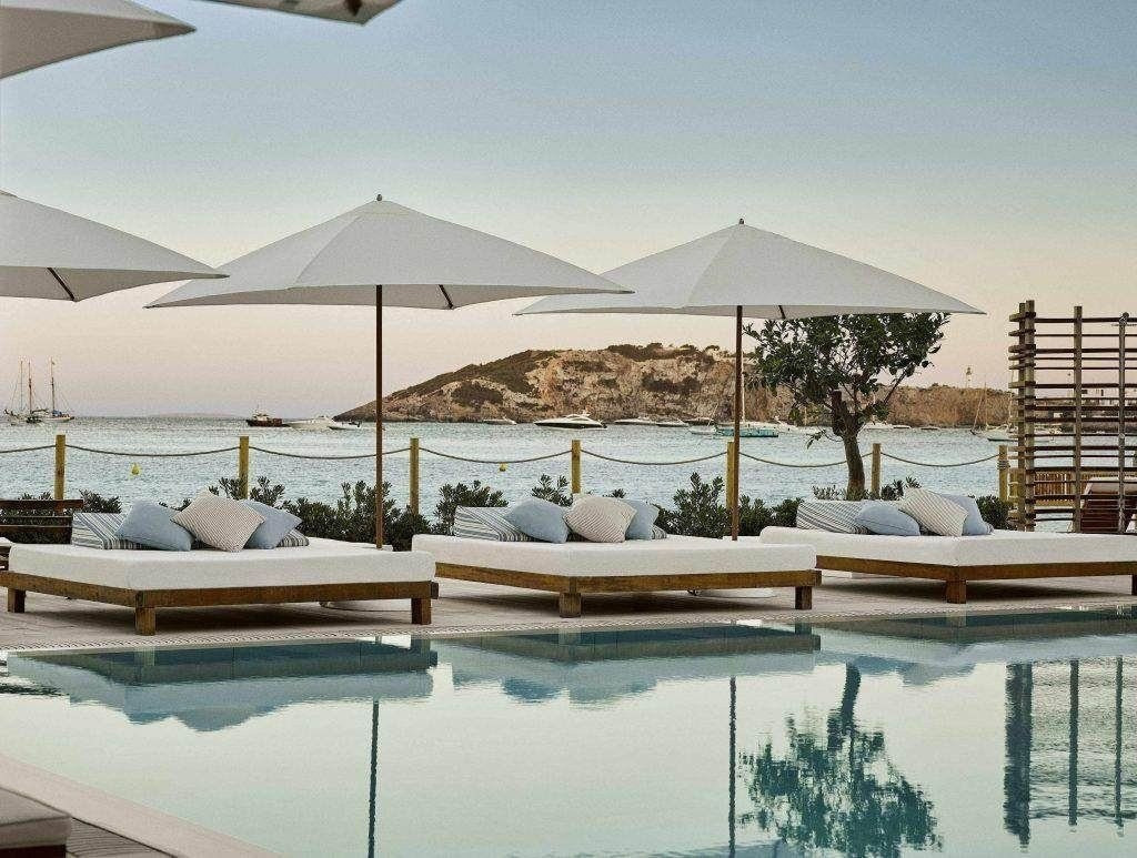 Drei luxuriöse, weiße Daybeds mit Sonnenschirmen stehen ordentlich aufgereiht an einem Pool mit Blick auf das ruhige Meer und ferne Hügel in der Abenddämmerung.