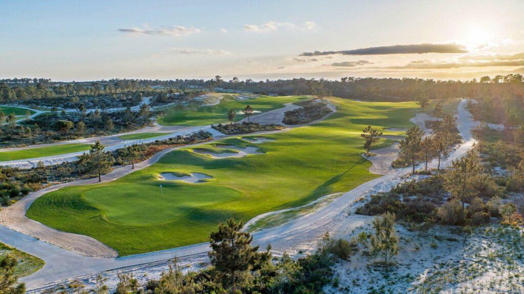 Eine Weitwinkelaufnahme eines Golfplatzes bei Sonnenuntergang. Die Szenerie zeigt wellige, leuchtend grüne Fairways, die von ausgedehnten, hellen Sandflächen (Waste Areas) und sandigen Wegen gesäumt sind. Vereinzelte Pinien werfen lange Schatten auf das Gelände. Im Hintergrund ist eine bewaldete Hügelkette unter einem klaren Himmel mit sanften Abendwolken zu sehen; die Sonne steht tief am rechten Bildrand und taucht die gesamte Landschaft in ein warmes, goldenes Licht.