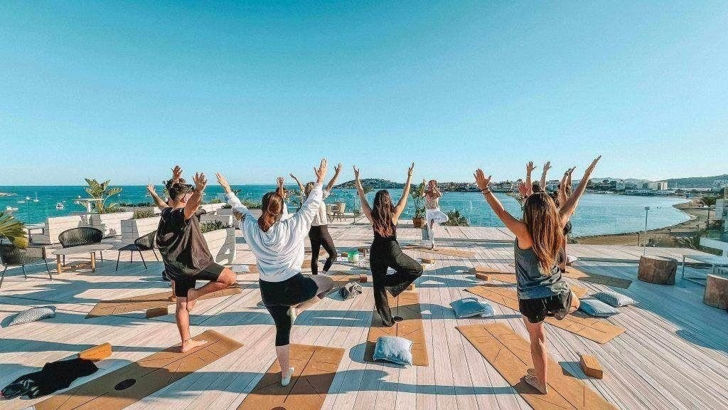Eine Gruppe von Menschen praktiziert Yoga in der Baum-Pose auf einem Holzdeck unter blauem Himmel mit direktem Blick auf das Meer.