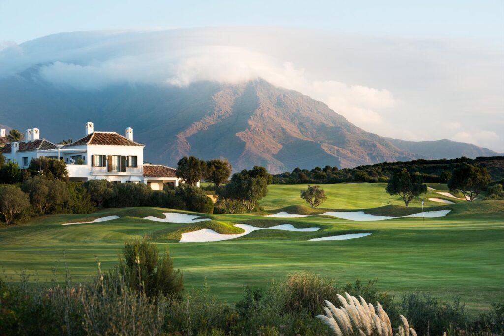 Ein saftig grüner Golfplatz mit weißen Sandbunkern im Vordergrund, eine weiße Villa im mediterranen Stil links und ein massiver Berg mit einer markanten Wolkendecke im Hintergrund.