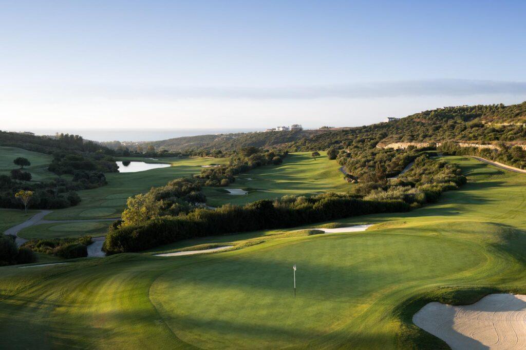 Hochperspektive auf ein langes Golf-Fairway mit einem Green und Flagge im Vordergrund, einem See auf der linken Seite und dem blauen Meer am fernen Horizont.