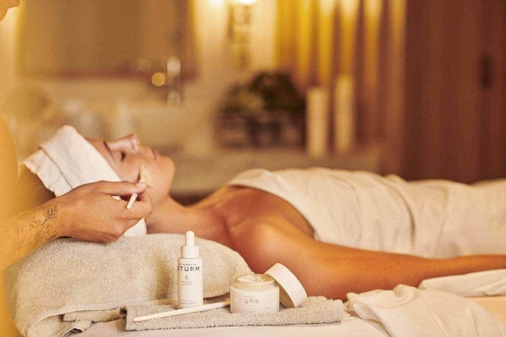 Eine Person liegt entspannt bei einer Gesichtsbehandlung in einem warm beleuchteten Spa; im Vordergrund stehen Pflegeprodukte von Dr. Barbara Sturm auf einem Handtuch.