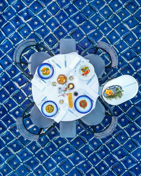 Ein runder Esstisch für vier Personen mit Speisen und Getränken, von oben gesehen, auf einem blau gemusterten Kachelboden.