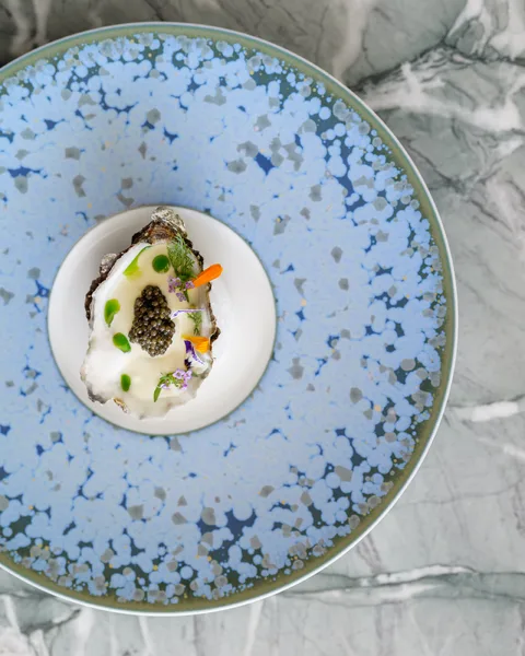 Eine Gourmet-Auster mit Kaviar-Topping und essbaren Blüten, serviert auf einem hellblauen, gesprenkelten Teller im Restaurant Pavyllon.
